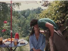 two people kissing under an umbrella on a patio table with food and drinks in front of them