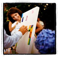 a woman is smiling as she paints on an easel while another person looks on