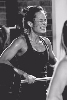 a woman is smiling while holding a barbell in front of her and another man behind her