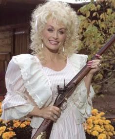 a woman holding two shotguns in her right hand and wearing a white dress with ruffled sleeves