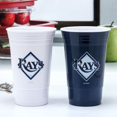 two plastic cups sitting next to each other on top of a white counter with fruit in the background