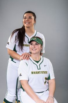 two female softball players are posing for a photo in their white uniforms and green caps