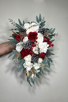 a bridal bouquet with red and white flowers, pine cones and greenery is being held by someone's hand