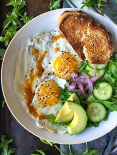 a white plate topped with an egg, bread and salad next to sliced cucumbers