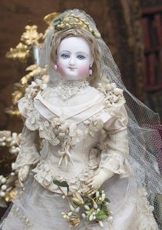 a doll is wearing a white dress and veil with flowers in her hair, sitting on a table