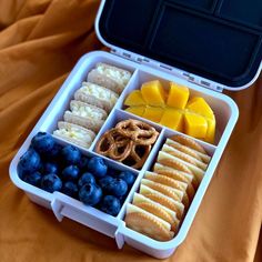 an open lunch box with fruit, crackers and pretzels in it on a bed