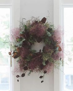 a wreath hanging on the side of a white wall next to a window filled with flowers