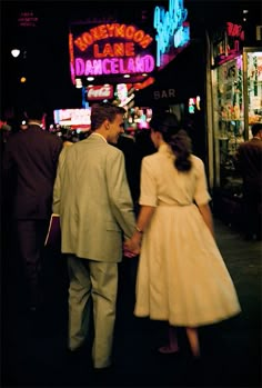 a man and woman holding hands walking down the street in front of a neon sign