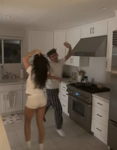 two people dancing in a kitchen with white cabinets and floor tiles on the walls,