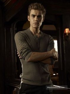 a man standing with his arms crossed wearing a sweater and black pants in a dark room