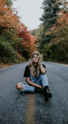 a woman sitting on the side of a road in front of trees with orange leaves