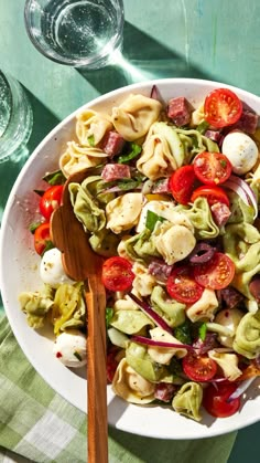 a white bowl filled with pasta salad next to two wine glasses and a wooden spoon