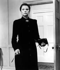 a woman in a long black coat and tie standing next to an open door with her hands on her hips