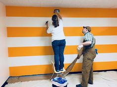 two people are painting a wall with orange and white stripes on it, while one man is using a paint roller