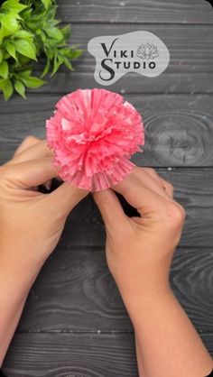 two hands holding a pink paper flower on top of a wooden table next to a potted plant