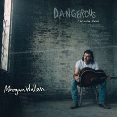 a man sitting on top of a chair holding an acoustic guitar in front of a brick wall