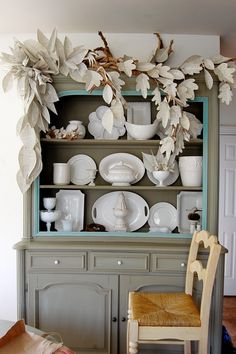 a dining room hutch with plates and cups on it's shelf, next to a chair