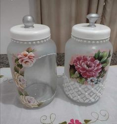 two glass jars with flowers painted on them sitting on top of a white table cloth