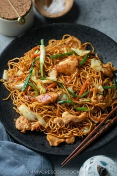 a black plate topped with chicken and noodles next to chopsticks on a table