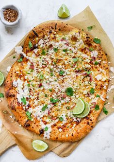 a pizza sitting on top of a wooden cutting board next to limes and seasoning