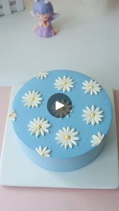 a blue cake with white daisies on it sitting on a table next to a figurine