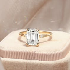 an emerald cut diamond ring sits on top of a white velvet box, with diamonds surrounding it