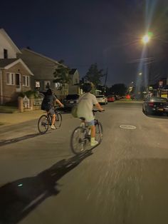 two people riding bikes down the middle of a street at night with cars behind them