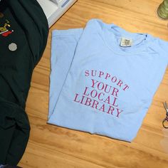 Show off your love for books and community with our "Support Your Local Library" embroidered shirt or crewneck. This cozy, customizable librarian sweatshirt is the perfect way to share your passion for reading and support the spaces that foster knowledge. Whether you're a proud book nerd or simply love curling up with a good read, this library lover tee makes a thoughtful gift for any literary enthusiast. Crafted for comfort and style, this bookworm t-shirt is perfect for anyone who values libra Library Shirt, Library Tshirt, White Bookish T-shirt As Gift, Literary Relaxed Fit Crew Neck T-shirt, Literary Graphic Print Crew Neck T-shirt, Bookish Crew Neck T-shirt With Graphic Print, Reading Day, Local Library, Gifts For Readers