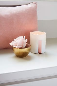 a white candle sitting on top of a table next to a pink pillow and a gold bowl