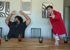 two men sitting at a table with drinks in front of them and one man holding his hands up