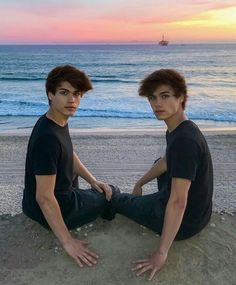 two young men sitting on the beach at sunset