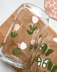 two glass vases with flowers cut out of them sitting on a wooden cutting board