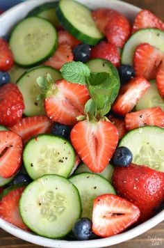 a bowl filled with cucumbers, strawberries and blueberries on top of a wooden table