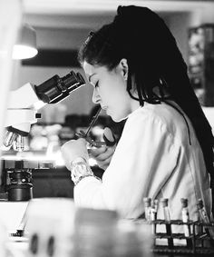 a woman looking through a microscope while sitting at a desk in front of a computer