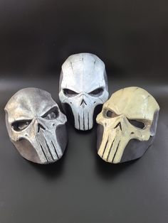 three different colored skull masks sitting on top of a black surface with white and gray paint