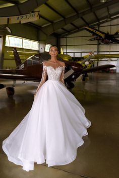 a woman standing in front of an airplane wearing a wedding dress with long sleeves and beading