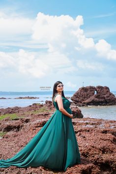 a woman in a long green dress standing on rocks near the ocean with her arms behind her back