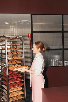 a woman in pink apron holding trays of doughnuts on metal rack at counter