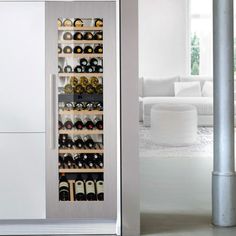 a wine cooler with many bottles in it next to a white couch and window on the wall