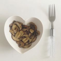 a heart shaped bowl filled with mushrooms next to a knife and fork on a white surface