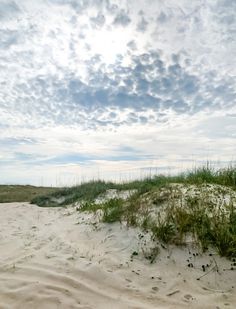 an empty sandy beach with grass growing out of the sand and clouds in the sky