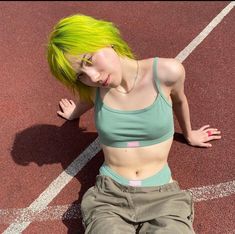 a woman with bright green hair sitting on a tennis court wearing khaki pants