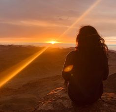 a woman sitting on top of a mountain watching the sun go down in the distance