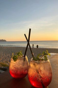 two glasses filled with drinks sitting on top of a wooden table next to the ocean