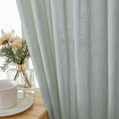 a cup and saucer sitting on a table next to a curtain with flowers in it