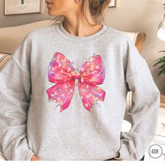 🎀 Coquette Pink Bow Christmas Sweatshirt. Features a pretty pink bow wrapped in twinkling Christmas lights--the ultimate vibe for anyone obsessed with all things coquette and cute. 💕✨ Wear this fun sweater to your next Christmas party or holiday movie night. Gift it to your BFF, your sis, or anyone who loves graphic shirts with a little holiday magic. 🎄 GET YOURS NOW ♡ H O W ∙ T O ∙ O R D E R * Select a Color * Select a Size based on the Size Chart * Add to Cart * Please check the Shipping Address to ensure that your order arrives without delay ♡ P R O D U C T∙ D E T A I L S * Brand: Gildan * Fabric: Medium-heavy weight. 50% cotton, 50% polyester. Ribbed knit collar. * Fit: Runs true to size. Please size up for an oversized look * Your shirts are carefully made with attention to small d Gift Jewelry With Pink Bow, Cute Pink Bow Jewelry Gift, Sweatshirt With Bows On Side, Trendy Crew Neck T-shirt With Bow, Pink Crew Neck T-shirt With Bow, Holiday Movie Night, Teen Girl Gifts, Holiday Movie, Holiday Sweater