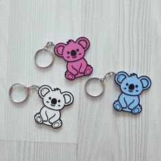 three key chains with different colored koalas and a teddy bear on them sitting on a wooden surface