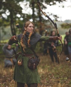 a woman dressed in medieval clothing holding an arrow and bow with other people behind her