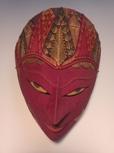 a red mask with yellow eyes on a white wall behind the mask is an intricate design