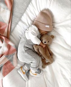 a small child laying on top of a bed next to a teddy bear wearing a hat